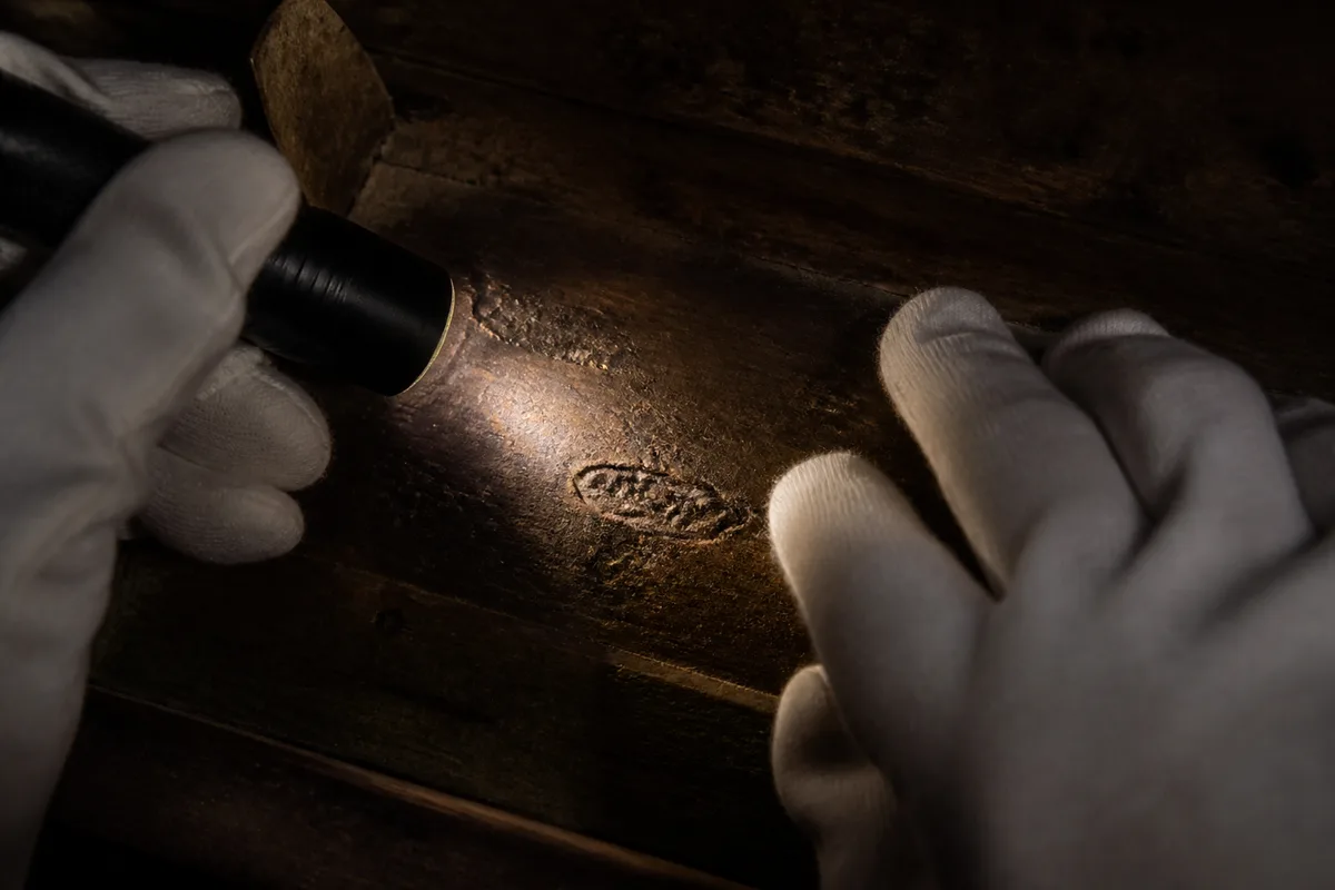 Mains gantées éclairant à la lampe torche une estampille frappée sur la traverse arrière d’une commode en bois sombre
