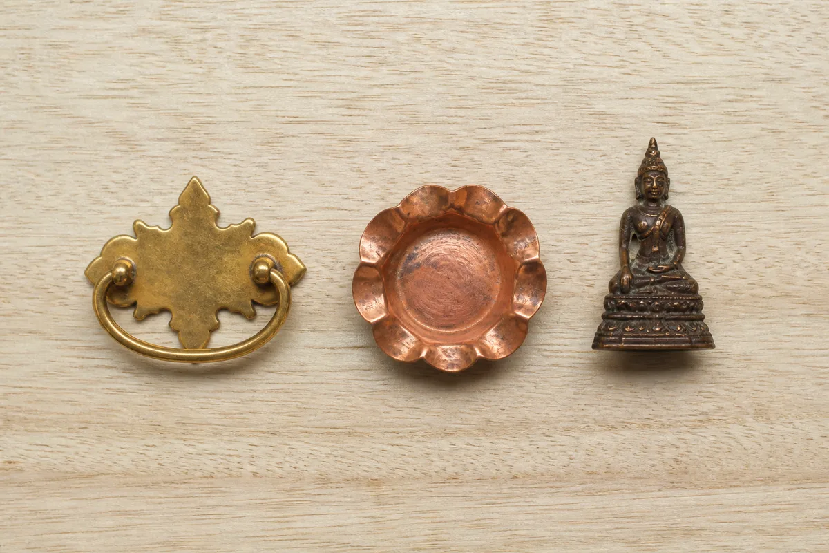 Poignée en laiton doré, coupelle en cuivre rosé et statuette en bronze sombre alignées sur un établi